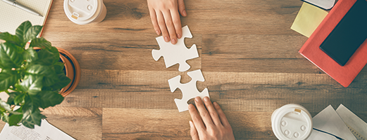 Zwei Hände setzen passende Puzzle-Teile auf einem Holztisch zusammen, als Symbol für Zusammenarbeit und Problemlösung.