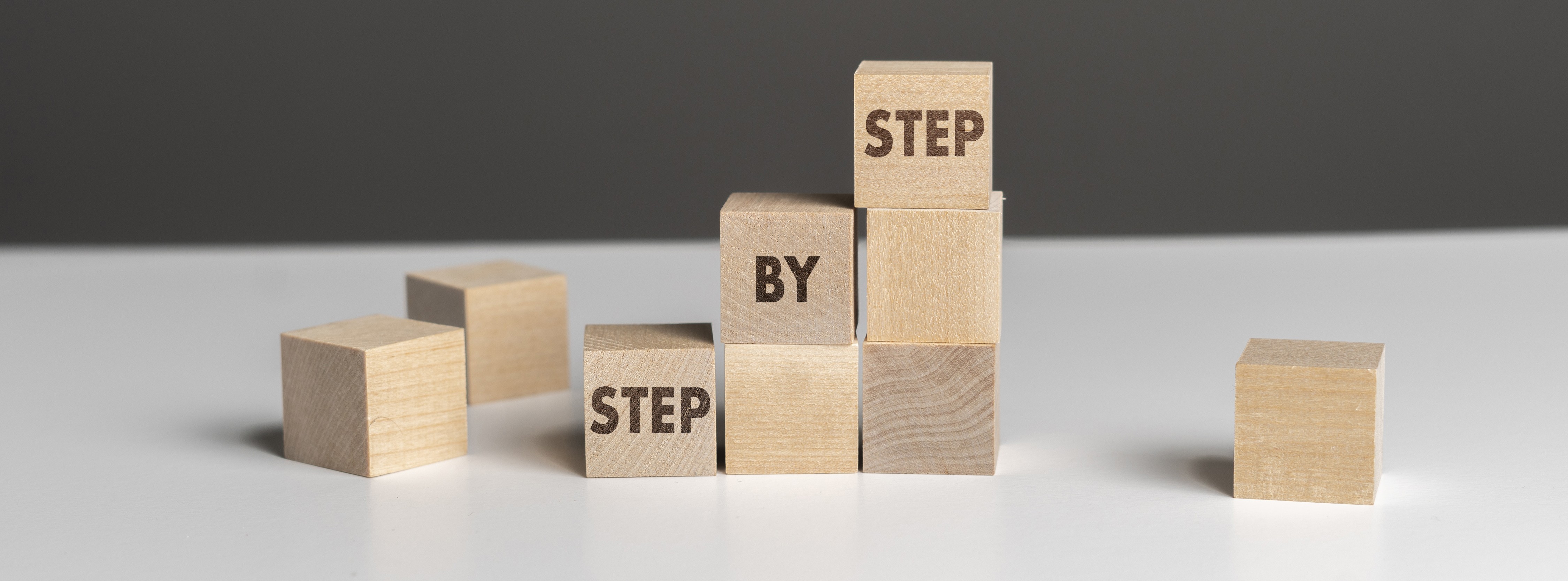Holzklötze bilden eine Treppe mit der Aufschrift ‚Step by Step‘ – Symbol für schrittweises Vorgehen.“