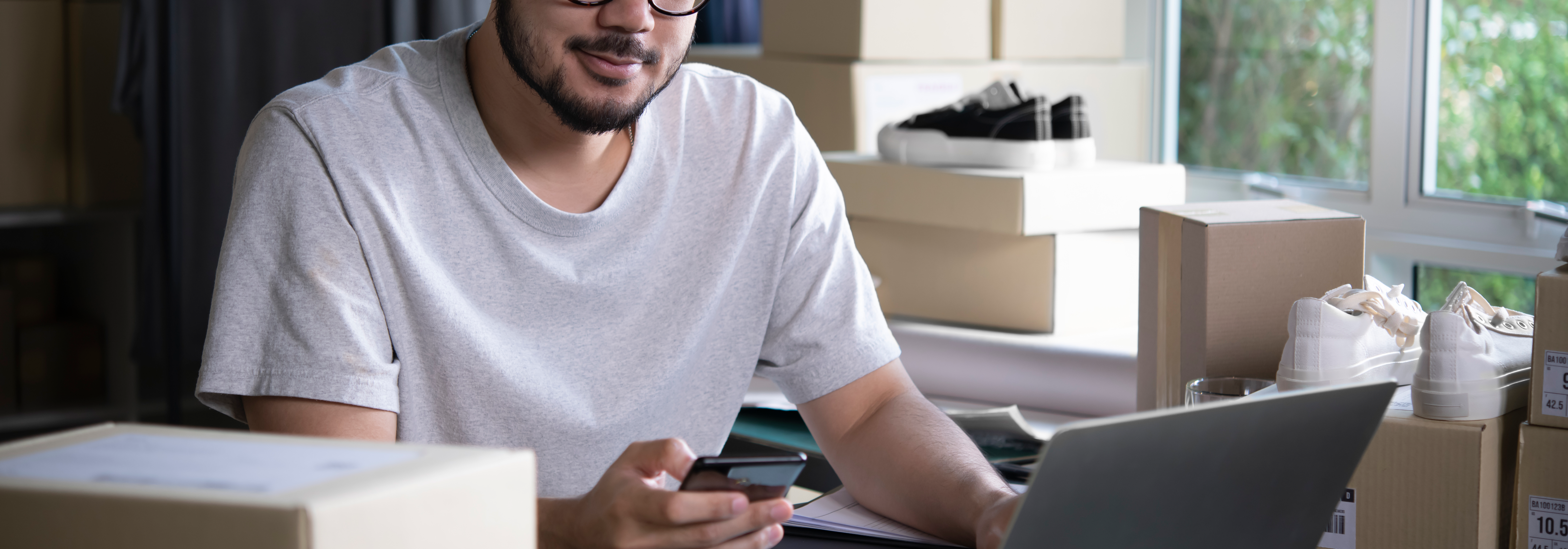 Mann arbeitet mit Laptop und Smartphone in einem kleinen Lager mit Schuhkartons – Onlinehändler prüft Bestellungen.